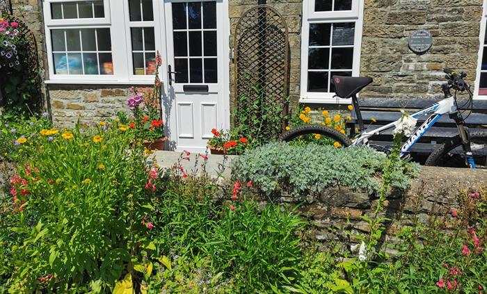 Close-up of a stone cottage entrance with a white door, colourful flower garden, and a bicycle resting against a low stone wall.